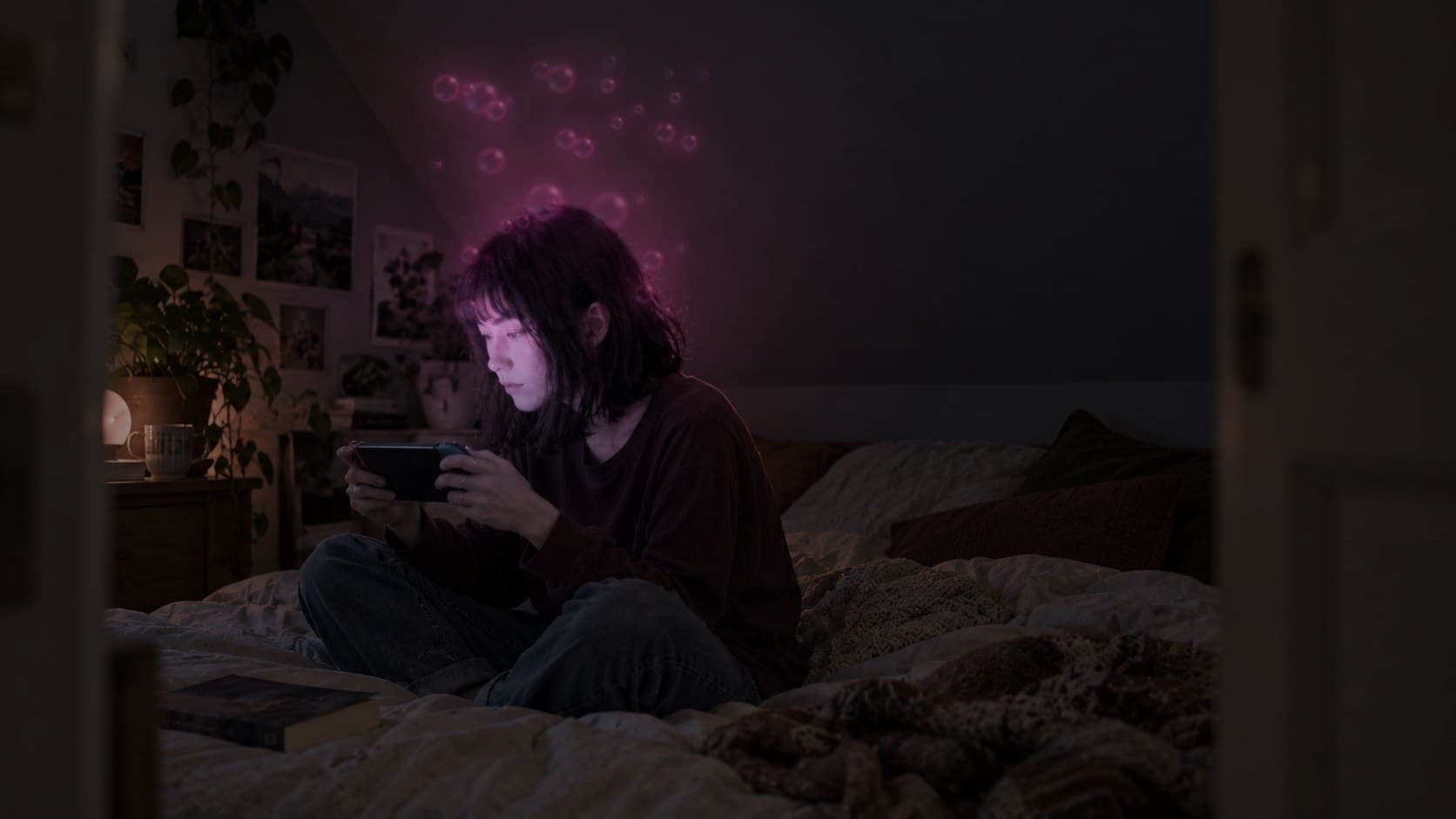 A woman with a black bob playing a handheld console in her attic bedroom, with softly glowing bubbles rising from her hair.